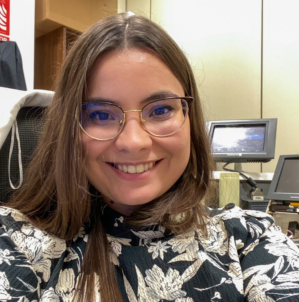 woman smiling at the camera wearing a black and white floral shirt and gold frame glasses sitting in front of computer equipment smiling at the camera
