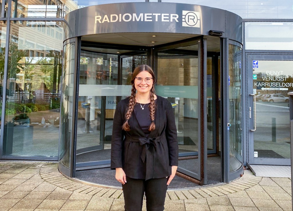 woman in all black outfit and braided pigtails standing in front of a rotating door with a sign above reading Radiometer