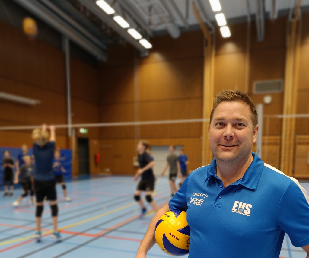 A man in a blue shirt holding a volleyball standing in front of other players and a volleyball net in the background.