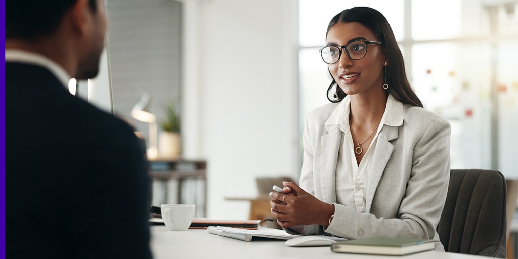 A woman in business casual outfit and glasses sits at a desk for a sales manager interview