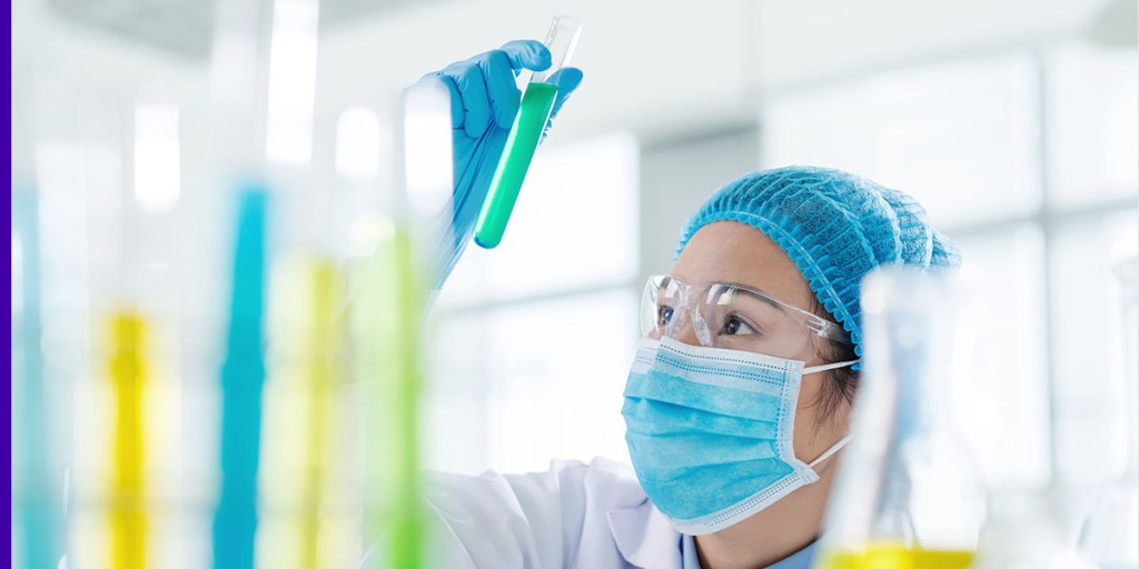 A woman in PPE looks at a test tube in a lab