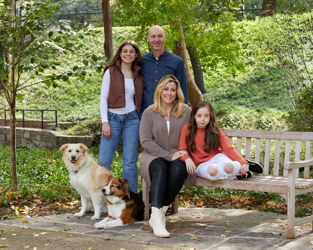 A family of four and two dogs sit on a bench together in a park.