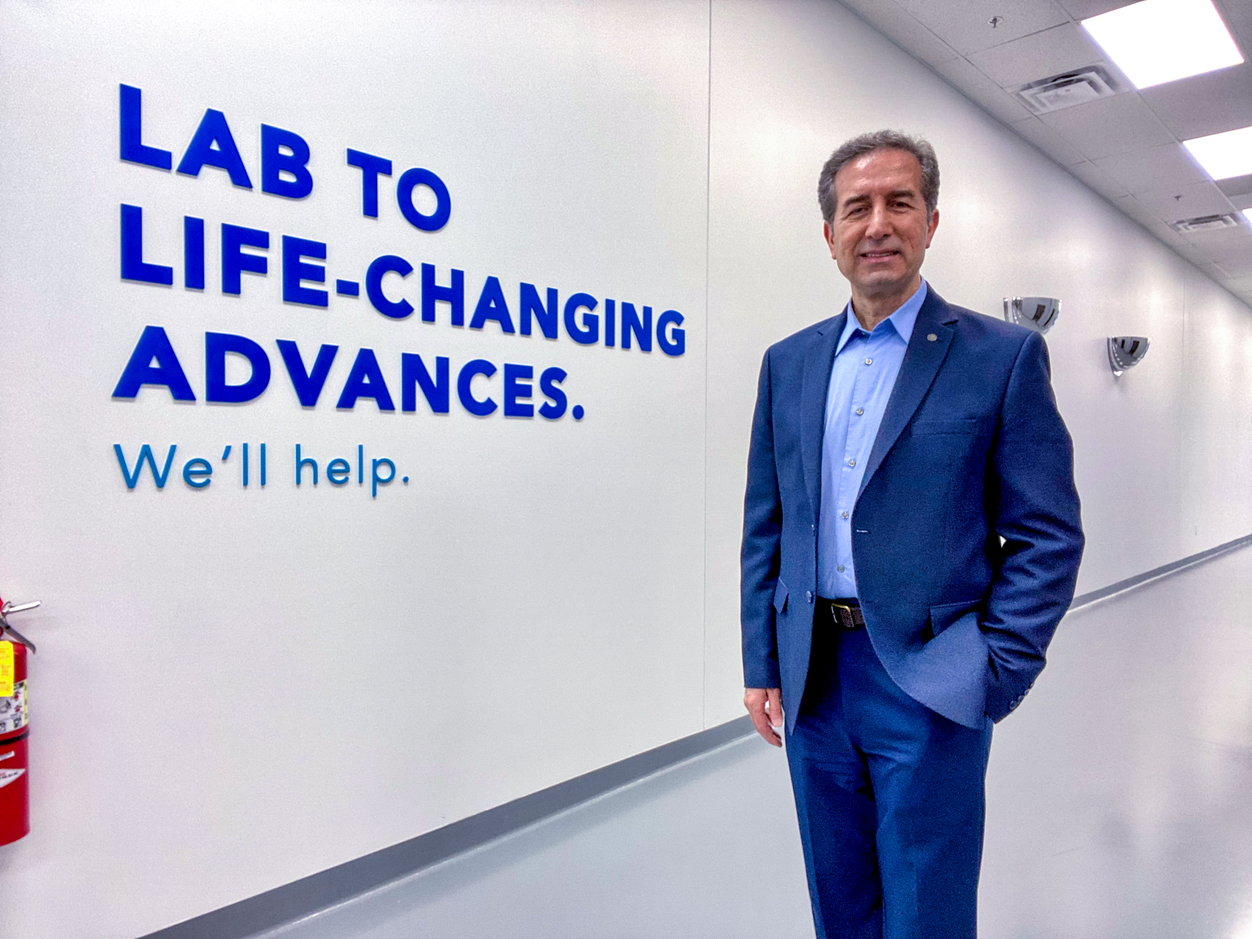 Dr. Karacay in a white hallway standing in front of a sign that says "Lab to Life-changing advances. We'll help."