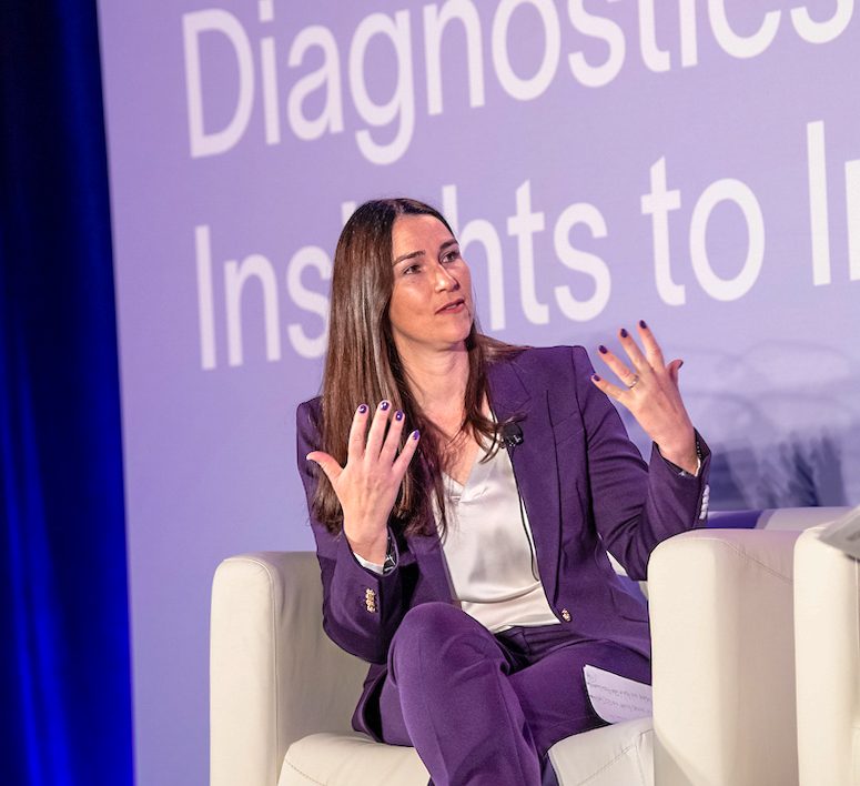 Two professionals in business attire engage in conversation at a Danaher Diagnostics event. They are standing in a modern conference space with branded signage and scientific displays in the background. Other attendees can be seen networking and viewing exhibits, creating a dynamic and professional atmosphere.