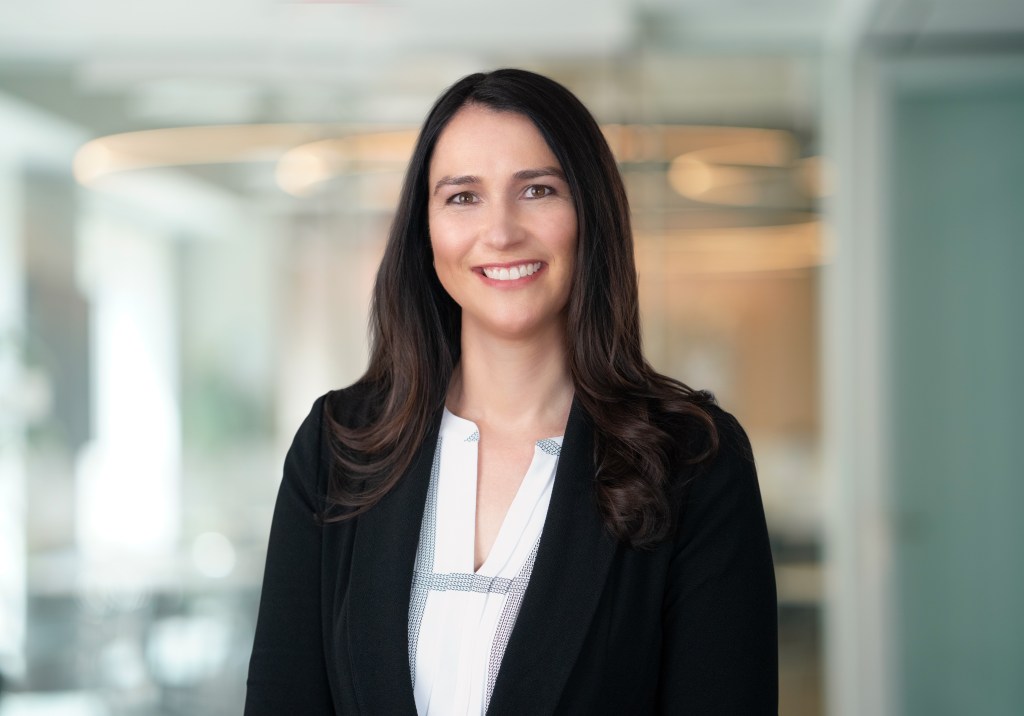 Headshot of a woman with long, straight dark brown hair parted to the side, smiling warmly. She is wearing a navy blazer over a white blouse. The background is softly blurred in neutral tones, keeping the focus on her face.