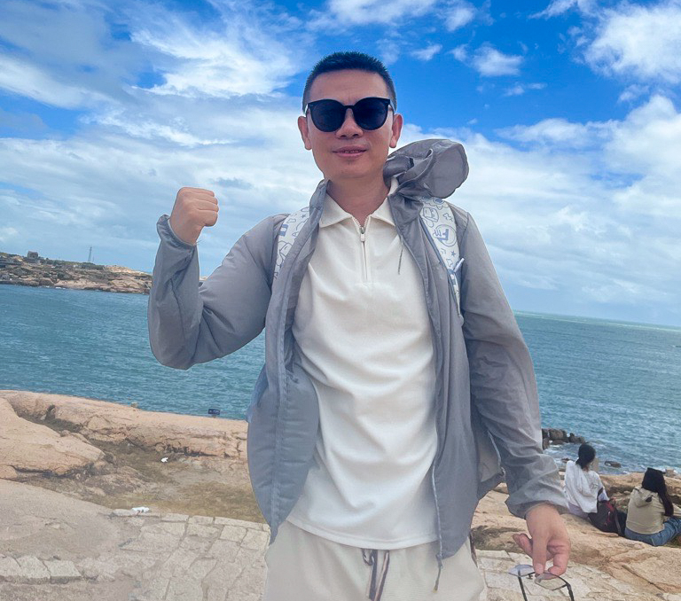 man wearing a white half zip shirt, khaki pants and a gray rain jacked, posing for the camera in front of a body of water