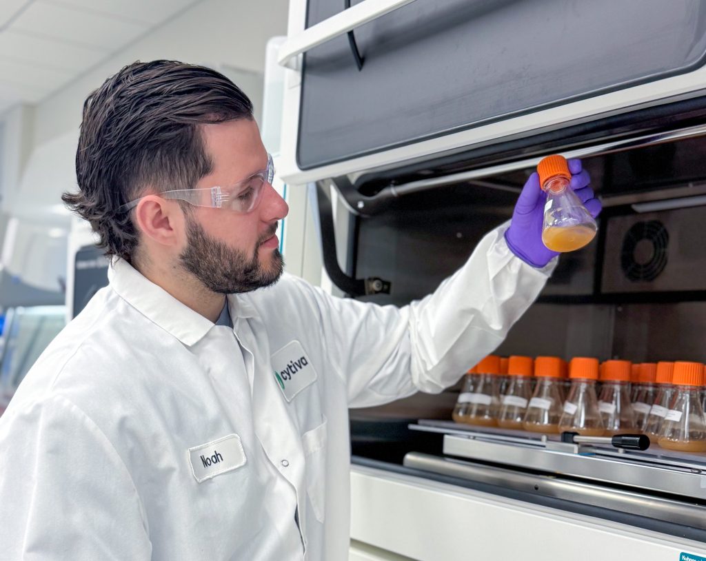 a man standing in a laboratory with gloves and a white coat on, examining a flask with an orange top