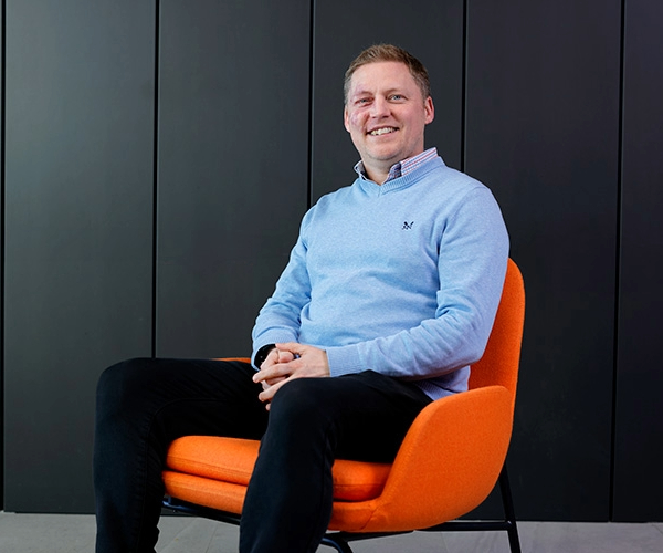 a man wearing a blue sweater and black pants sitting in an orange upholstered chair and posing for the camera