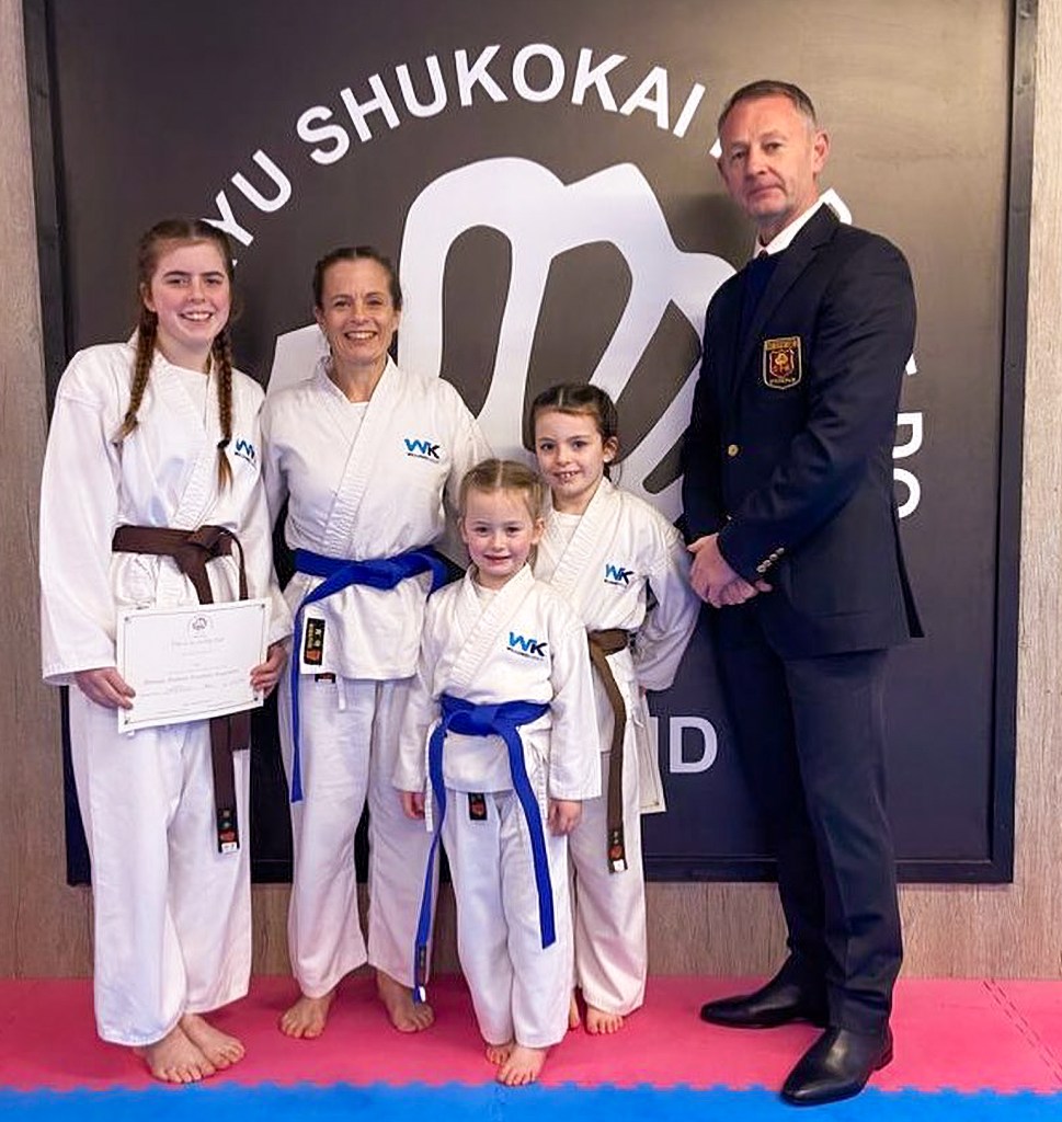 a woman and three young girls in karate uniforms post in front of a sign alongside an older man