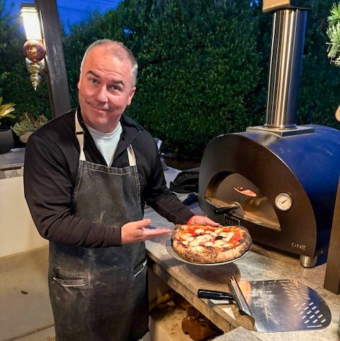matt hicks holding a pizza in front of a tabletop pizza oven in an outdoor kitchen
