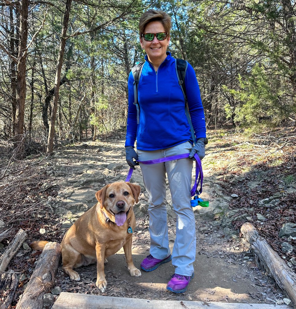 Jessica standing on a wooded trail with a yellow labrador sitting to her left