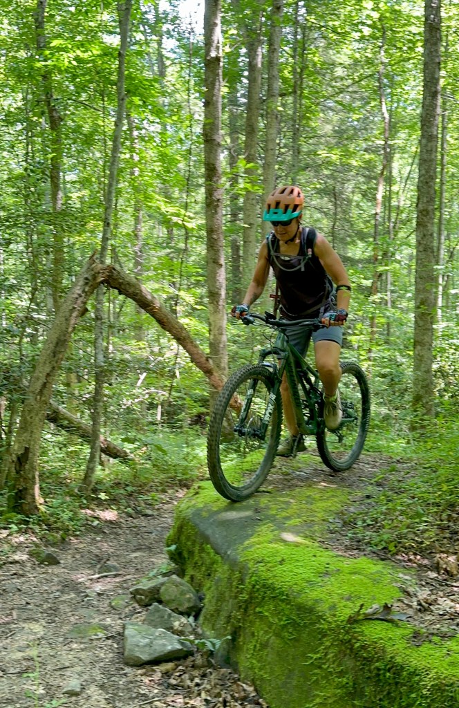 Jessica riding a bike through a wooded trail down a steep step