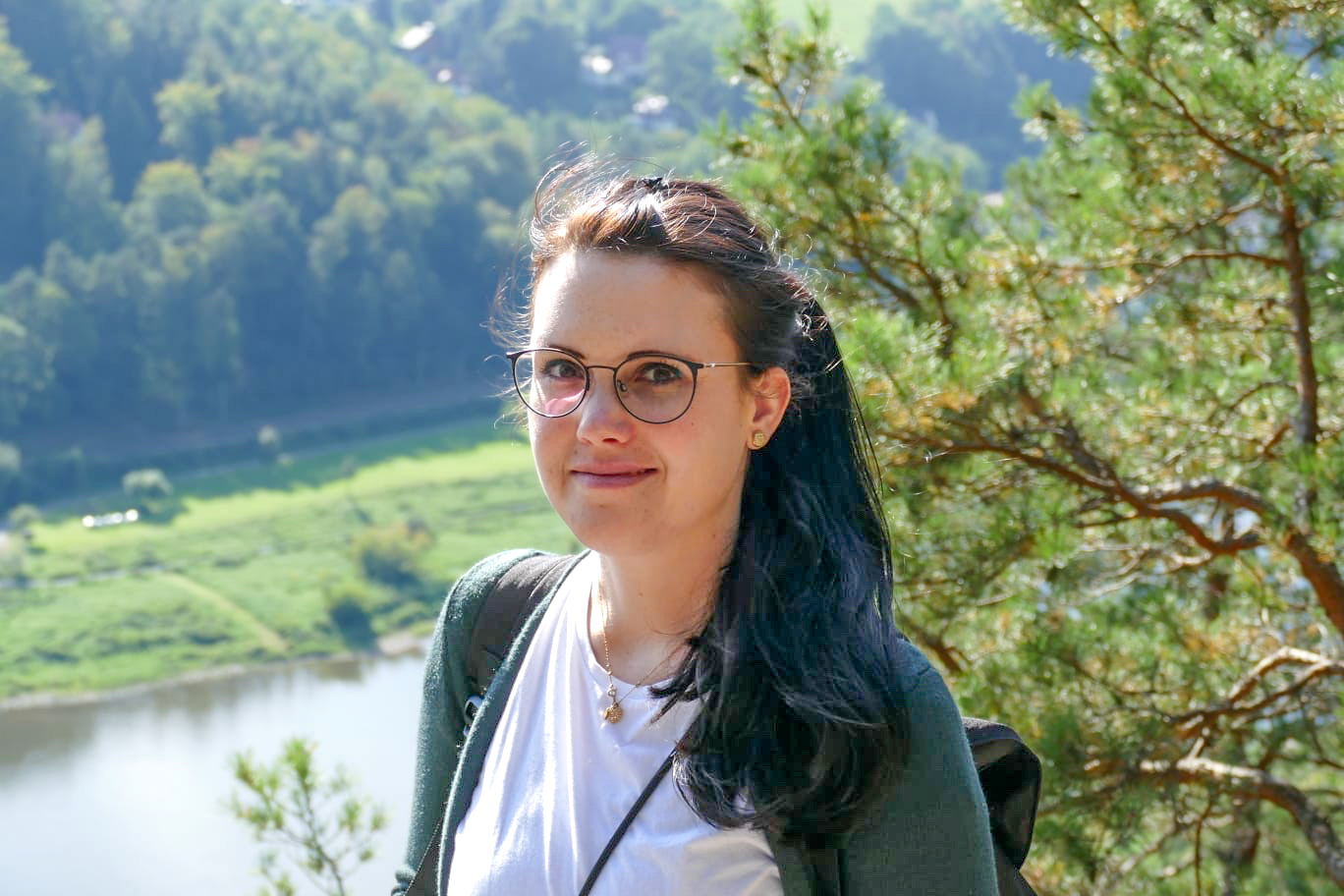 Person wearing glasses and outdoor clothing standing near a river and trees on a sunny day.