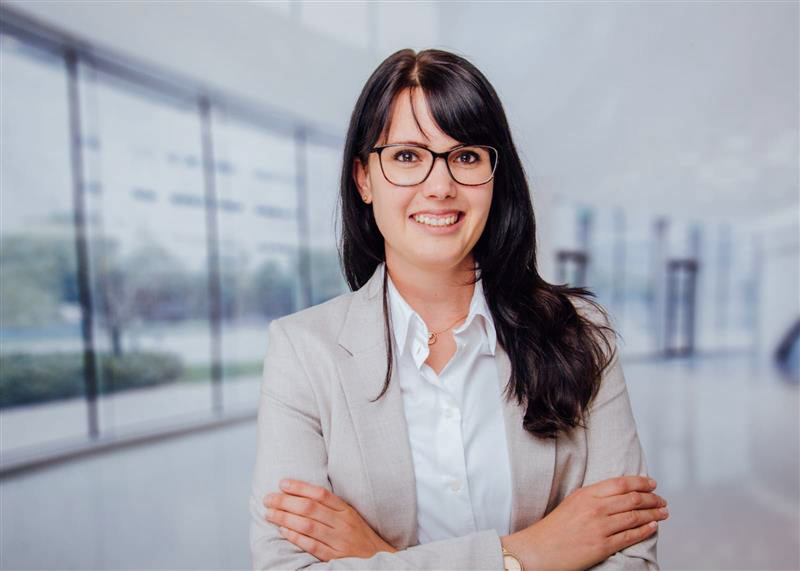 Professional portrait of a person wearing business attire, standing indoors with arms crossed in a modern office setting.